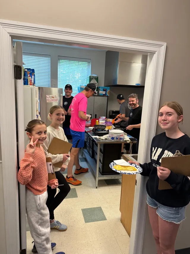 Three girls with clipboards smile in a kitchen doorway, one holding an omelet on a paper plate, as men cook behind them.