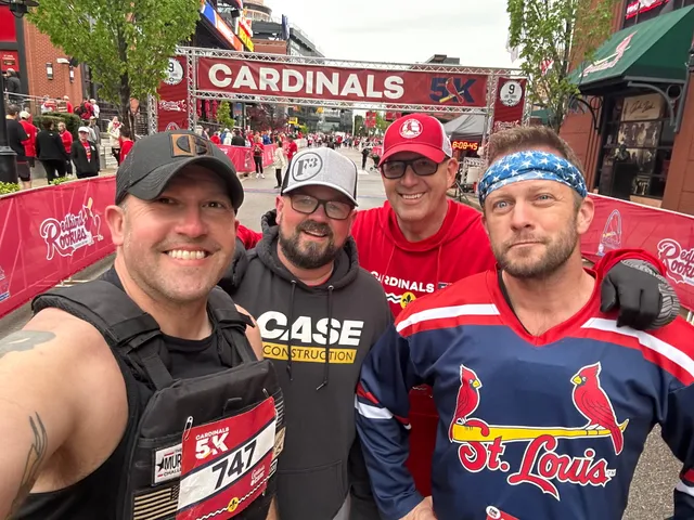 Four men smile for a selfie in front of the Cardinals 5K finish line arch, one wearing a weighted vest and race bib 747.