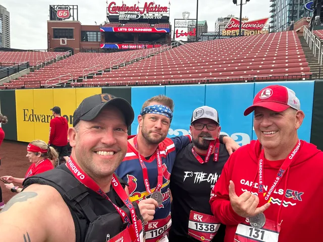 Four smiling men wearing race medals and bibs take a selfie on the field at a baseball stadium with red seats behind them