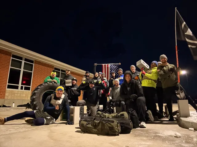 Group of men in winter hats pose at night outside a brick building with a large tire, cinder blocks, rucksacks and a US flag