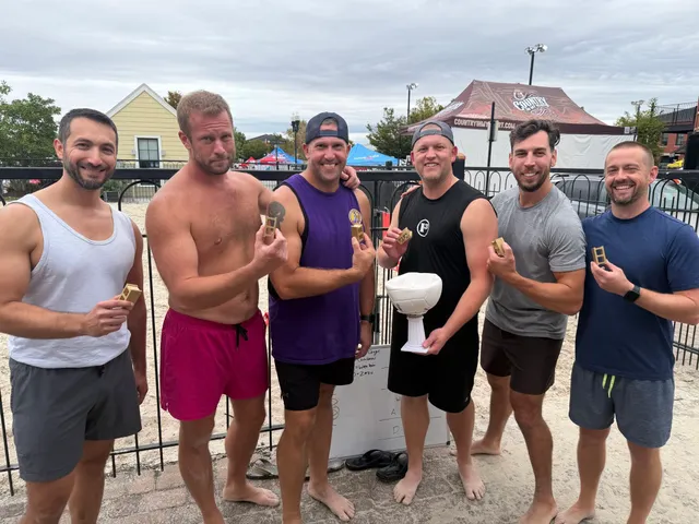 Six barefoot men in tank tops and shorts on a sand court hold small gold trophies; one holds a white volleyball trophy.