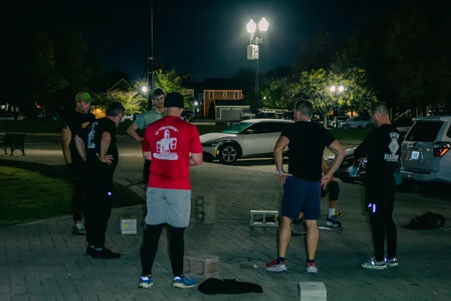 Men in workout clothes stand in a circle on a lit brick plaza at night, cinder blocks on the ground near parked cars.