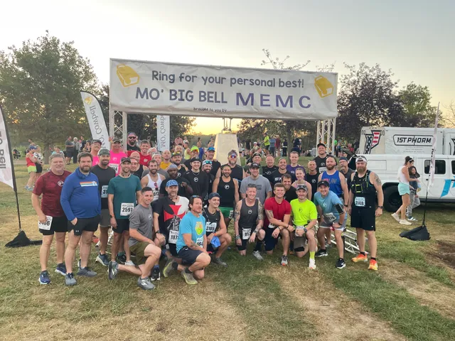 Large group of runners with race bibs posing under a "Mo' Big Bell MEMC" banner at a race finish area at dusk
