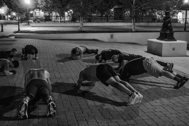 Black-and-white photo of nine men doing push-ups and planks on a brick plaza at night under streetlights.