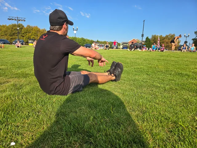 Man in black cap and shirt sits on grass stretching toward his toes while a group sits in a wide circle on a sunny lawn