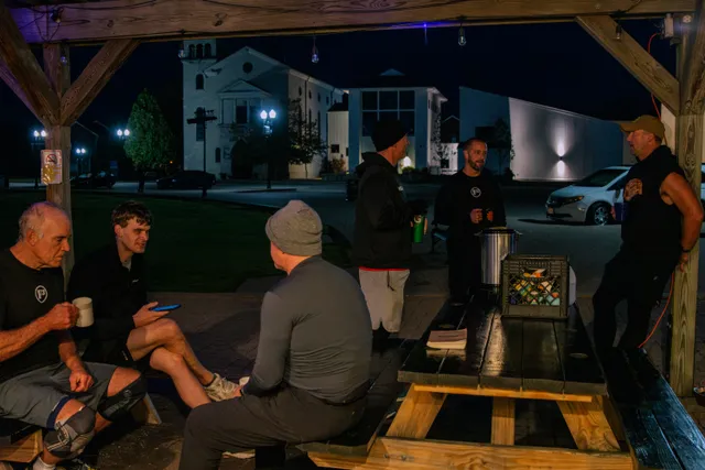 Men in workout clothes chat with mugs around a wooden picnic table under a lit pavilion at night, buildings behind.