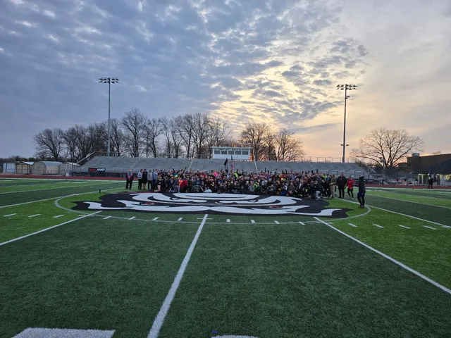 Large group poses on a football field's midfield logo at dawn, with empty bleachers, bare trees and stadium lights behind