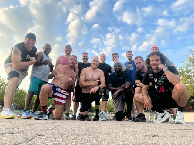 Twelve men in workout clothes pose on pavement under a cloudy blue sky, some kneeling, one in stars-and-stripes shorts.