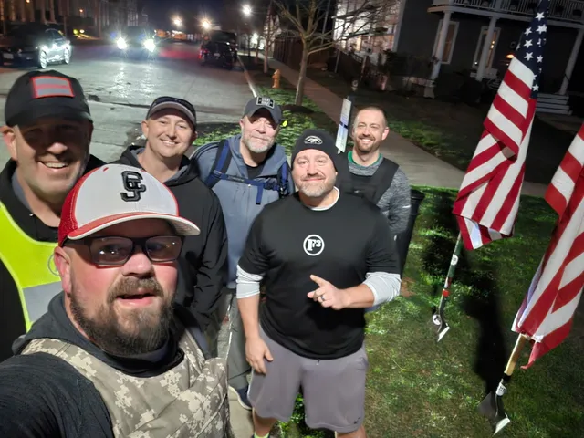 Six men pose for a nighttime selfie on a sidewalk beside two American flags mounted on shovels, wearing caps and weight vests.