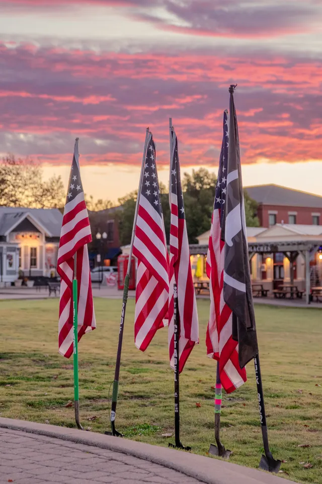 Four American flags and a black POW/MIA-style flag on shovel handles planted in grass under a pink sunset sky
