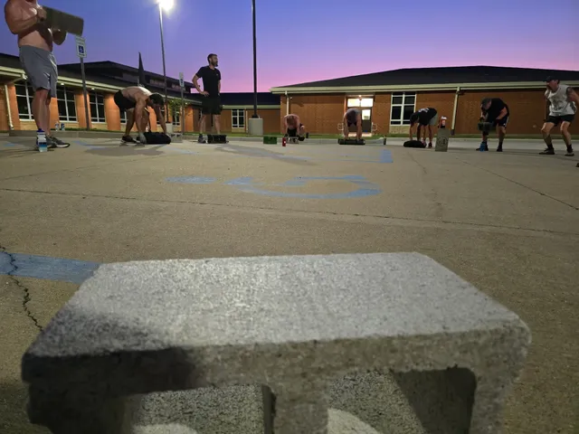 Low view past a cinder block of men exercising with sandbags and blocks in a parking lot at dawn under a pink sky