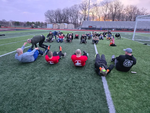 Large group of men doing sit-ups in a circle on a turf football field at dawn, with bleachers and bare trees behind
