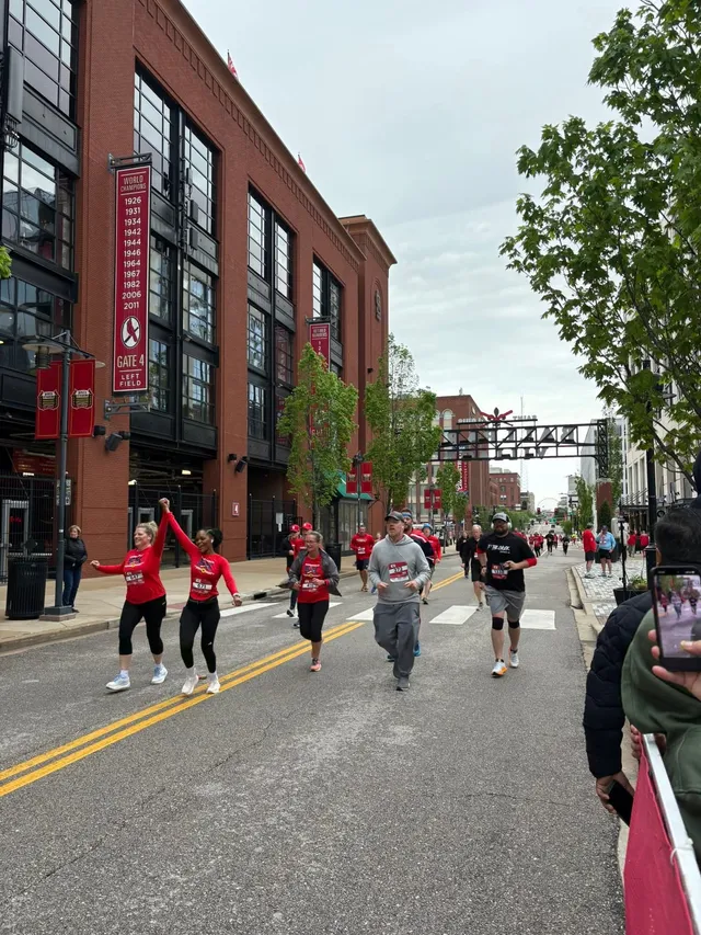Runners with race bibs, many in red shirts, jog down a street past a brick stadium with a Gate 4 Left Field banner.