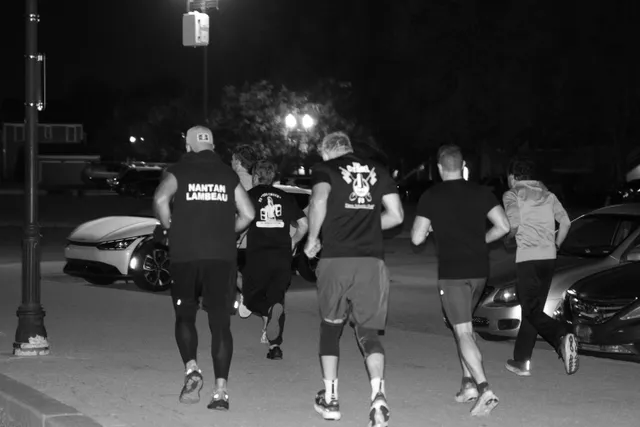 Black-and-white photo of a group of men running away from the camera at night past parked cars and streetlights.