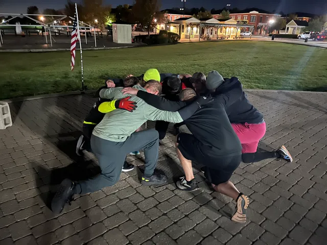 Group of men in athletic wear kneel in a huddle with arms around each other on brick pavers at night beside an American flag.