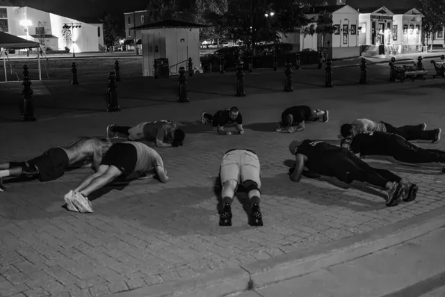 Black-and-white photo of about nine men holding plank and push-up positions on a brick plaza before dawn, bollards behind them