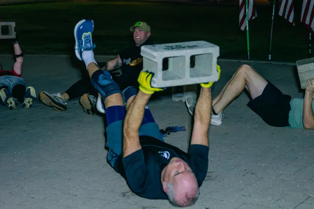 Man lying on pavement lifts a cinder block overhead with yellow gloves as others do the same in a predawn workout