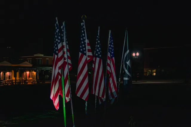 Five American flags and a dark green flag with a white logo on poles, lit in darkness before lamplit buildings