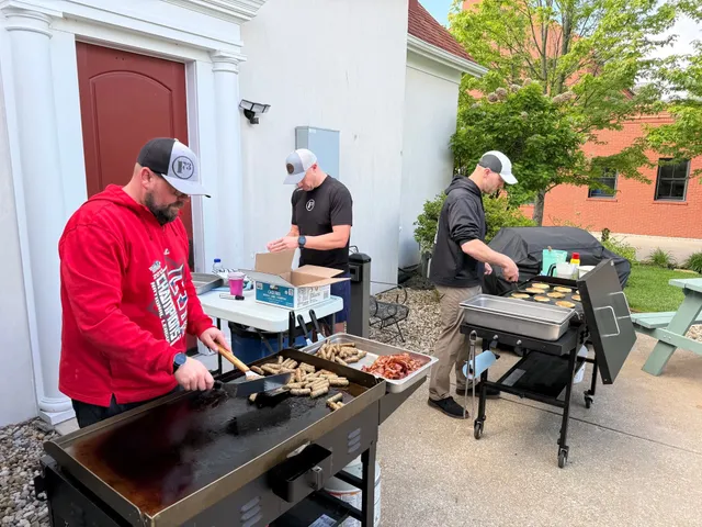 Three men in caps cook sausage, bacon and pancakes on outdoor flat-top griddles beside a white building.
