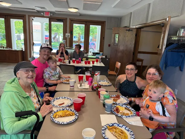 Family smiling at a breakfast table with omelets, bacon and red cups in a community room; a toddler sits on a woman's lap.