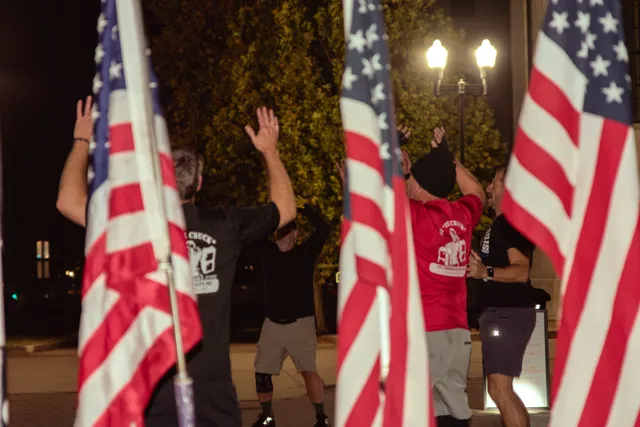 Men in workout clothes raise their arms in a circle at night, framed by American flags under a lit streetlamp.