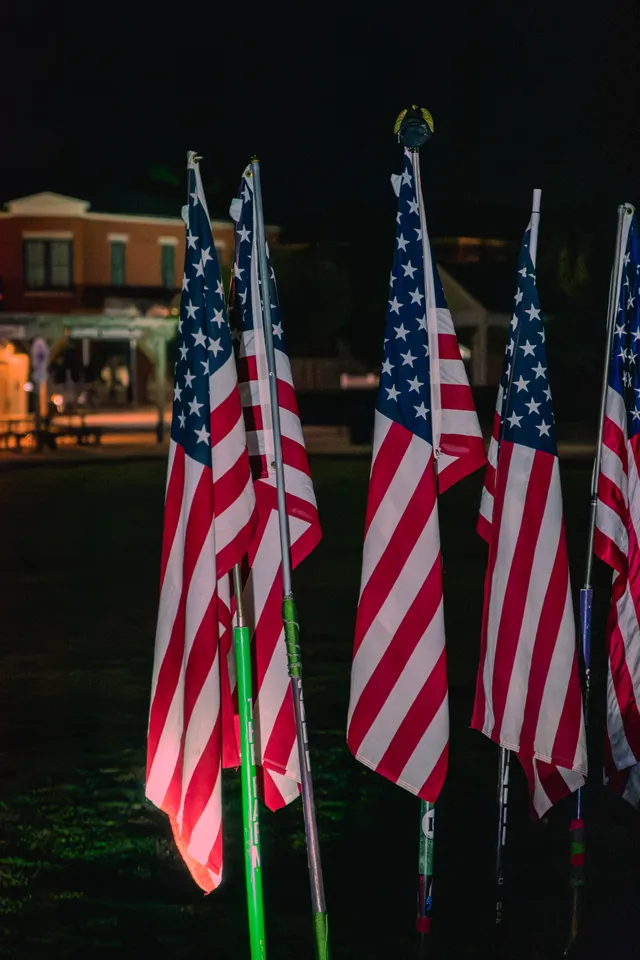 Five American flags on poles, some taped to hockey sticks, standing on dark grass at night with a lit brick building behind