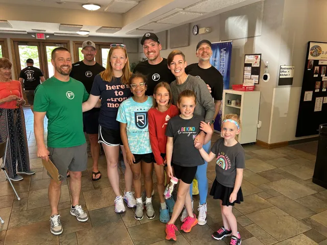 Group of adults in F3 shirts and four smiling girls pose in a tiled church lobby with a worship banner behind them.