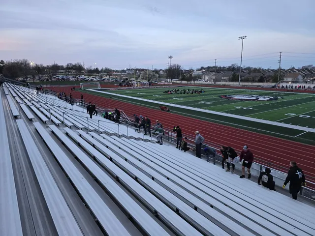 People exercising along the lower rows of aluminum stadium bleachers at dawn, with a group working out on the football field beyond.