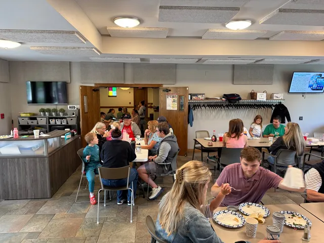 Families eat breakfast at tables in a community room with a coat rack, wall TVs and a serving counter.