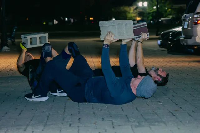 Three men lie on brick pavement at night pressing cinder blocks and a brick overhead, with parked cars behind them.