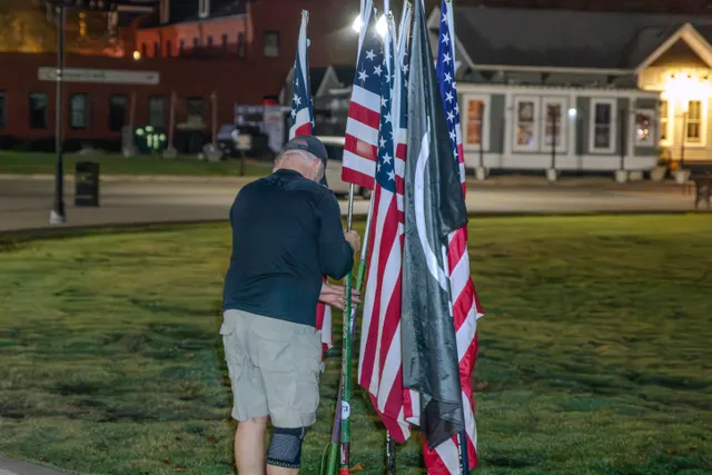 Man in dark shirt, cap and khaki shorts handles a cluster of American flags and a black POW/MIA flag on a lawn at night