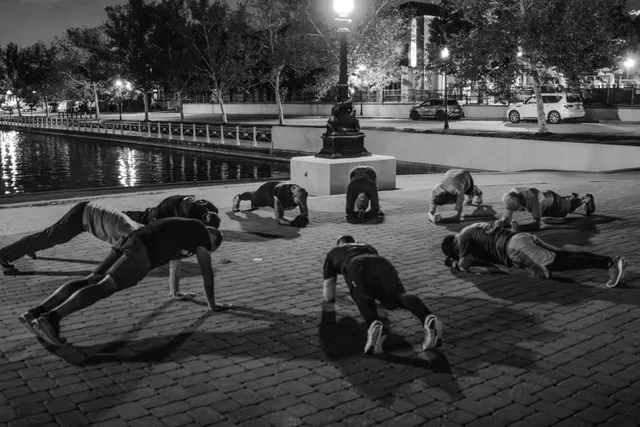 Black-and-white photo of men doing push-ups and planks on brick pavers beside a lit canal and lamppost before dawn