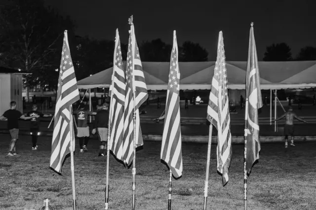 Black-and-white photo of six flags on poles in grass at dawn as men walk and stretch near a large white tent behind them.