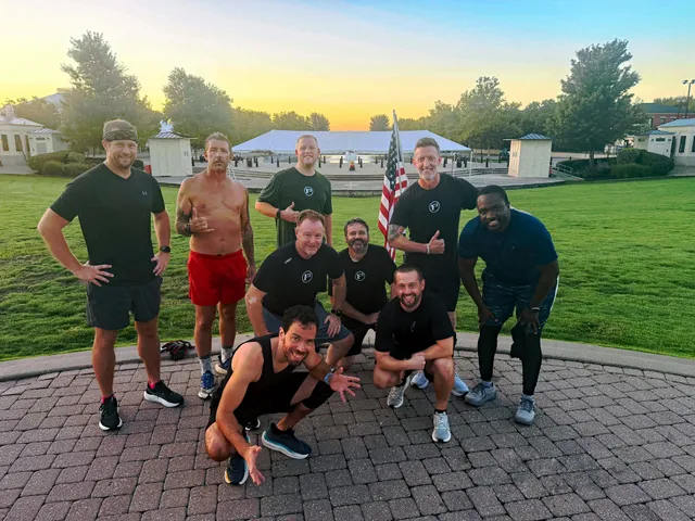 Nine men in workout clothes pose on a brick plaza at sunrise with an American flag, lawn and white tent behind them.