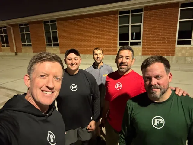 Five smiling men in F3 logo shirts pose for a nighttime selfie outside a red brick building with large windows.