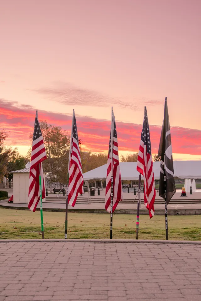 Four American flags and a black POW/MIA flag on labeled poles in a lawn at sunrise, white tent and fountain behind