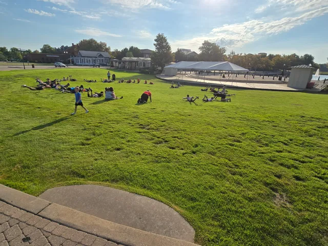 Adults and kids lie on a grassy hillside doing exercises near a white tent and amphitheater on a sunny evening