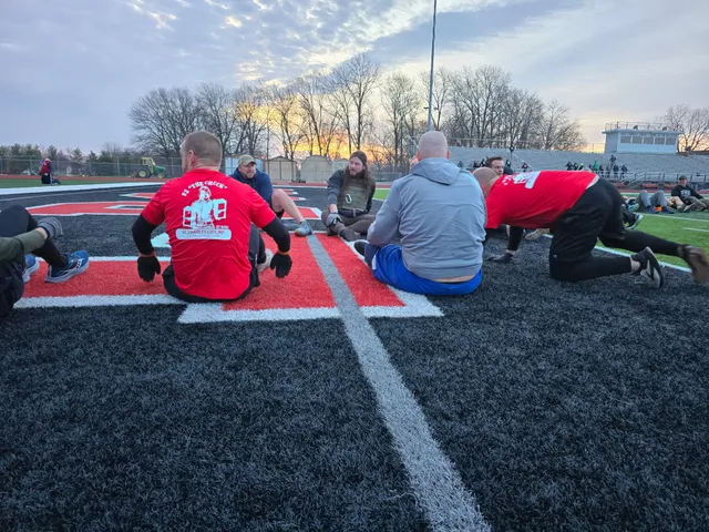 Men sit and stretch in a circle on a black turf football field with a red end zone logo at sunrise, bleachers behind.