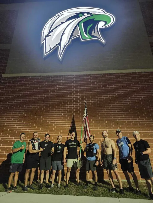 Nine men in workout clothes pose at night by a brick wall under a lit eagle logo, one holding a U.S. flag on a green pole