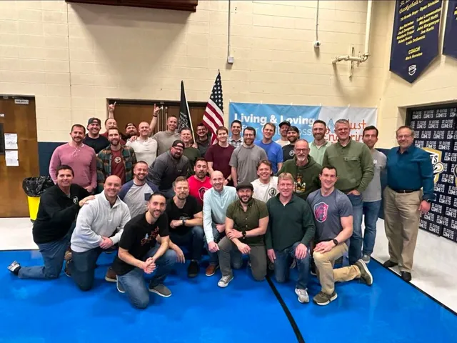 About 30 men pose in a school gym on a blue floor, some kneeling, with an American flag and a black flag behind them.