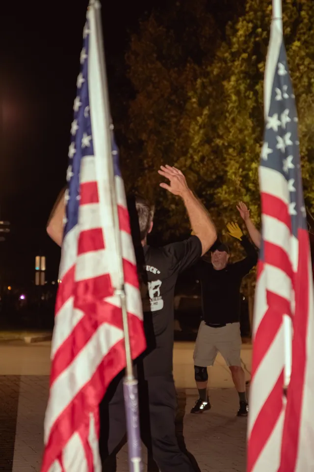 Two men in black shirts raise their arms during a nighttime outdoor workout, framed by two American flags in the foreground.
