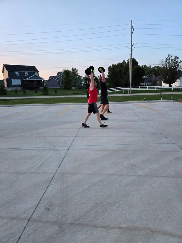 Men in shorts and gloves walk across an empty concrete parking lot holding black sandbags overhead at dawn