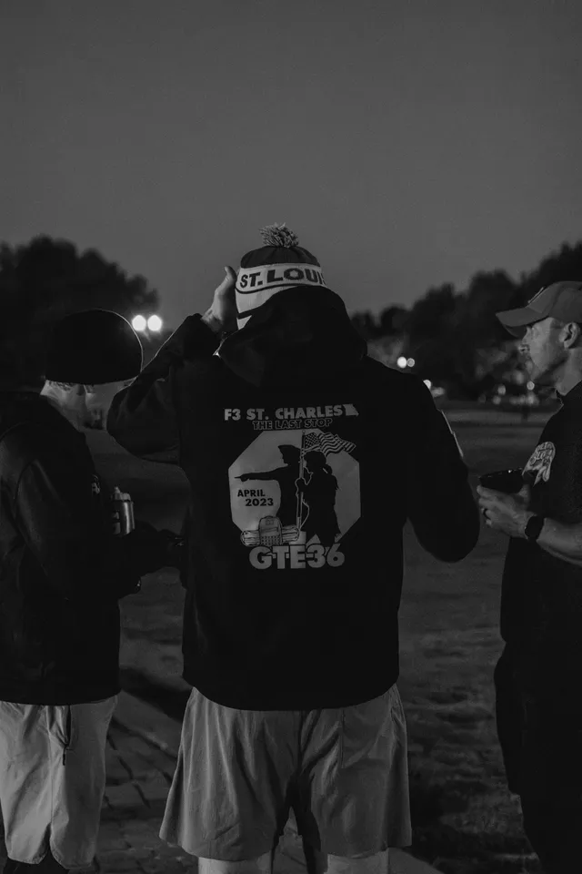 Black-and-white photo of a man from behind in an F3 St. Charles GTE36 hoodie and St. Louis beanie, standing with two men at dusk