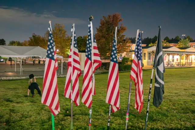 Five American flags and a dark green flag on poles in a grassy lawn at night as two men exercise behind them near a white tent