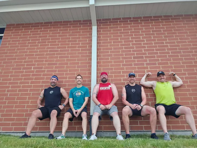 Five men in workout clothes do wall sits against a red brick wall above grass, one flexing both arms.