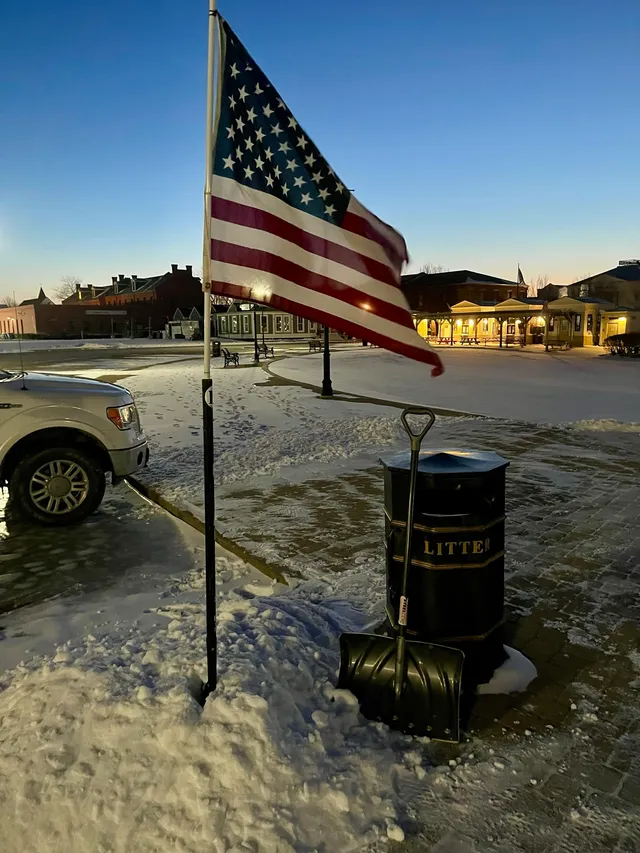 American flag on a pole beside a snow shovel leaning on a black LITTER bin in a snowy plaza at dawn, lit buildings behind