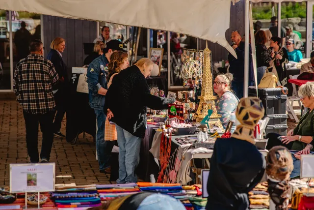 Shoppers browse vendor tables of jewelry, a gold Eiffel Tower display and colorful folded textiles under a white tent