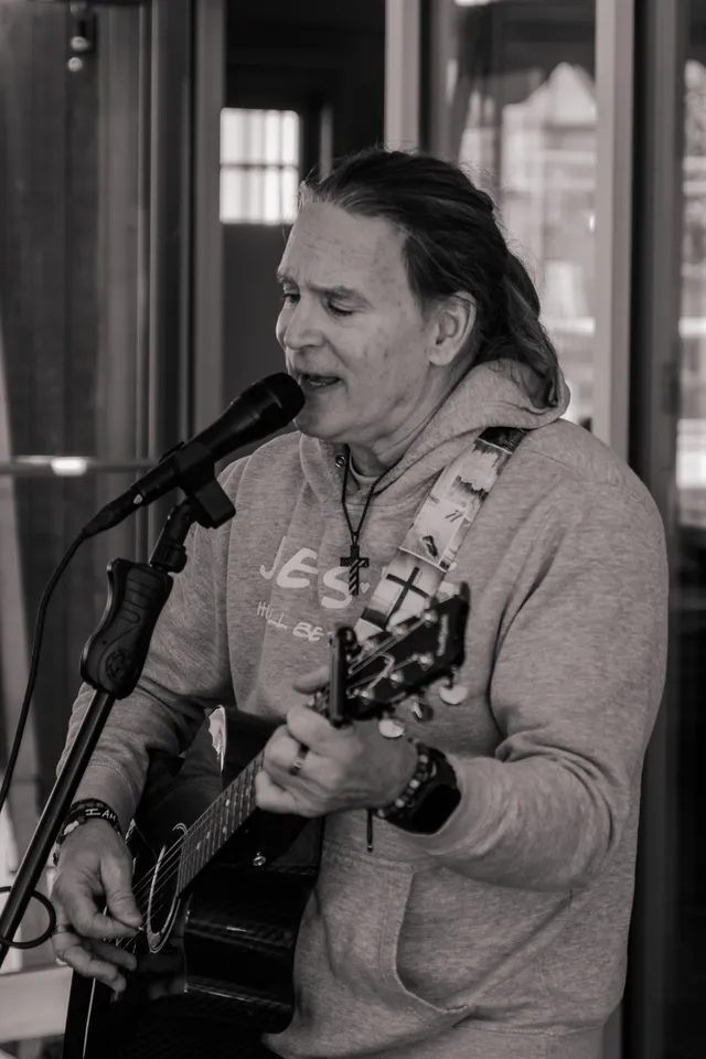 Black-and-white photo of a long-haired man singing into a microphone while playing a dark acoustic guitar