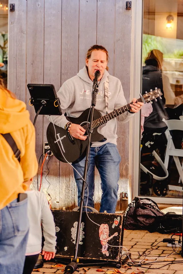 Musician in gray hoodie sings into a mic while playing a black acoustic guitar with a nail cross design, small child watching