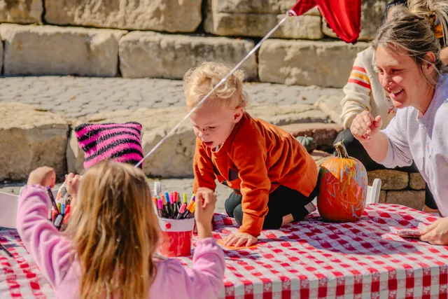 Toddler in orange sweater crawls on a red gingham table beside a marker-decorated pumpkin as a smiling woman watches.